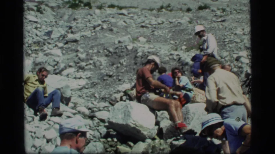 1977 Glacier Bay home-movie frame showing hikers seated and crouched among loose rocks in a mountain valley.