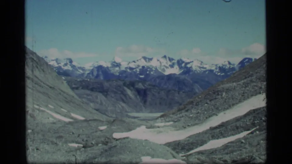 1977 Glacier Bay frame with a rocky glacial valley, lingering snow patches, blue sky, and distant snow-capped peaks.