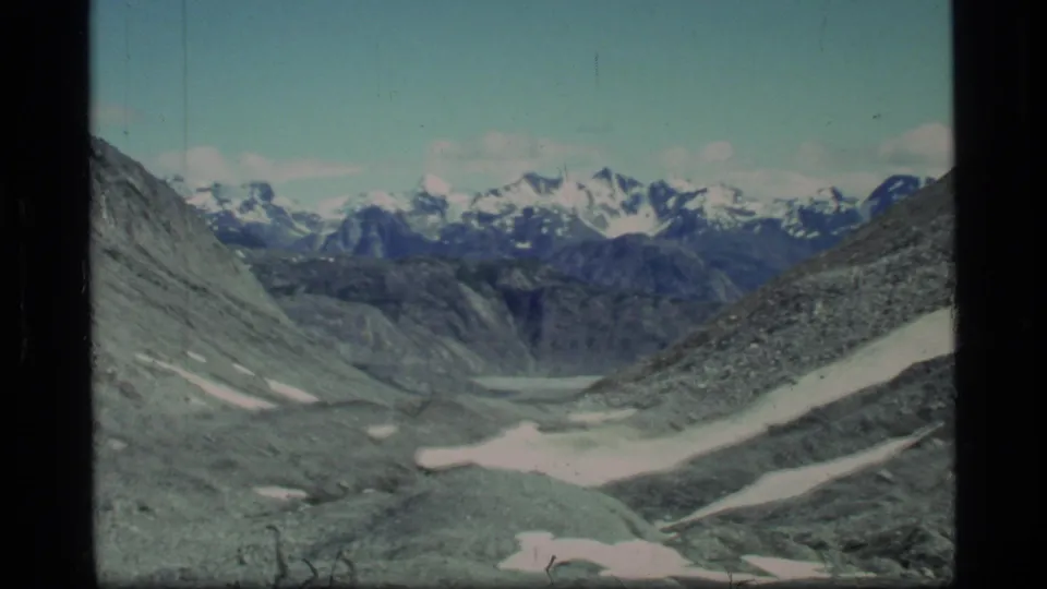 1977 Glacier Bay frame showing a broad rocky valley, white snow patches, and distant snow-capped peaks beneath clear sky.