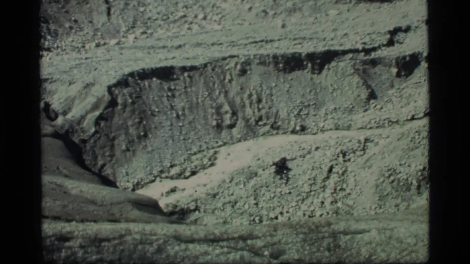 1977 Glacier Bay frame with light-toned mountain terrain, rough layers, and dark fissures across the surface.