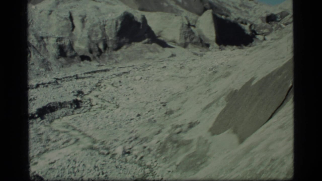 Glacier Bay landscape frame showing a debris-covered valley, steep pale slope, and dark rock outcrops under clear sky.