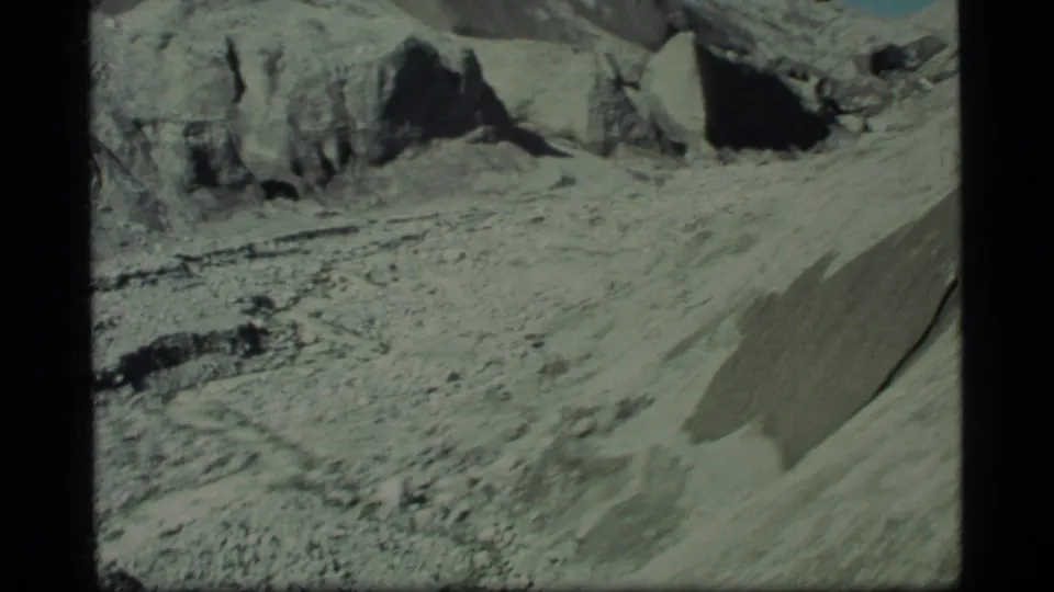 Glacier Bay landscape frame showing a debris-covered valley, steep pale slope, and dark rock outcrops under clear sky.