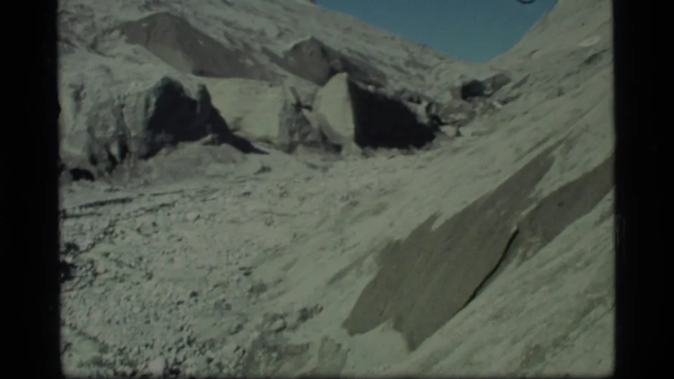 1977 Glacier Bay 8mm frame with large boulders, eroded mountain slopes, gravel foreground, and blue sky above the valley.