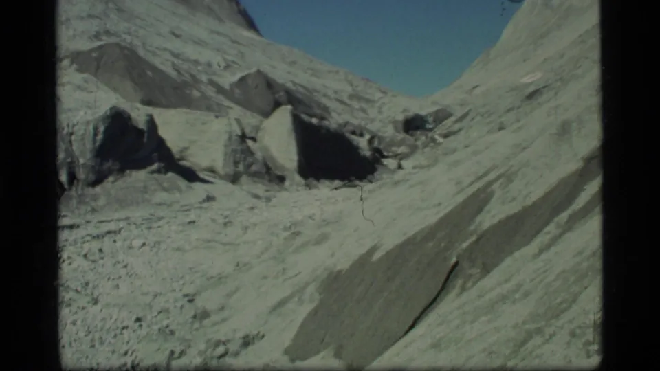 Wide 8mm Glacier Bay landscape view with boulders, pale gravel, steep slopes, and a small opening of clear blue sky.