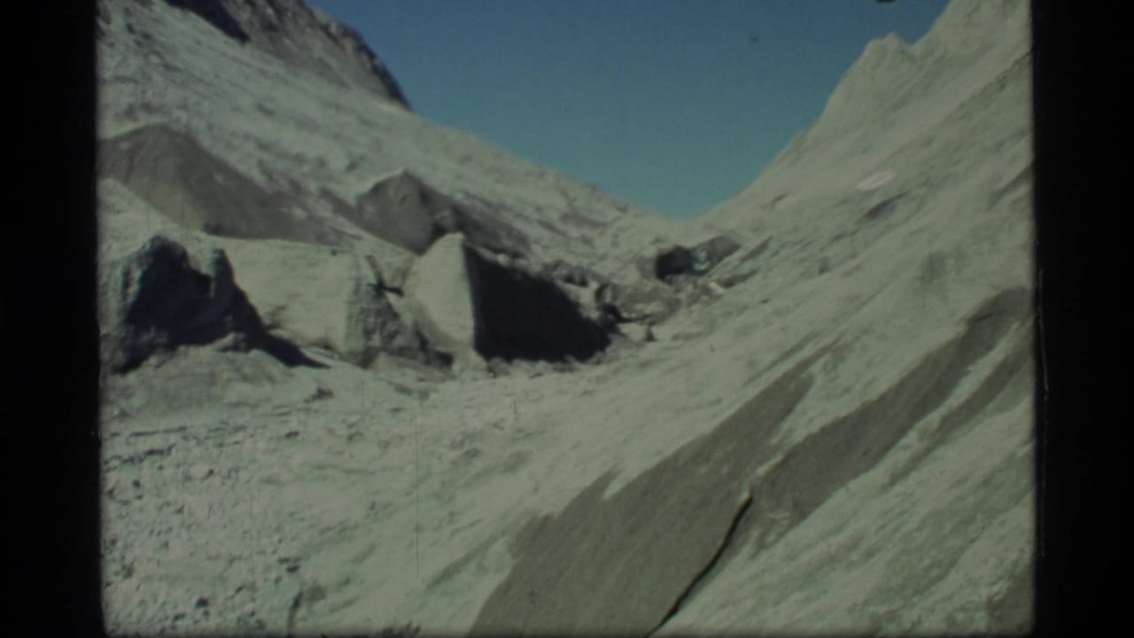 1977 8mm Glacier Bay view of pale rocky glacial terrain, scattered boulders, and steep slopes beneath clear blue sky.