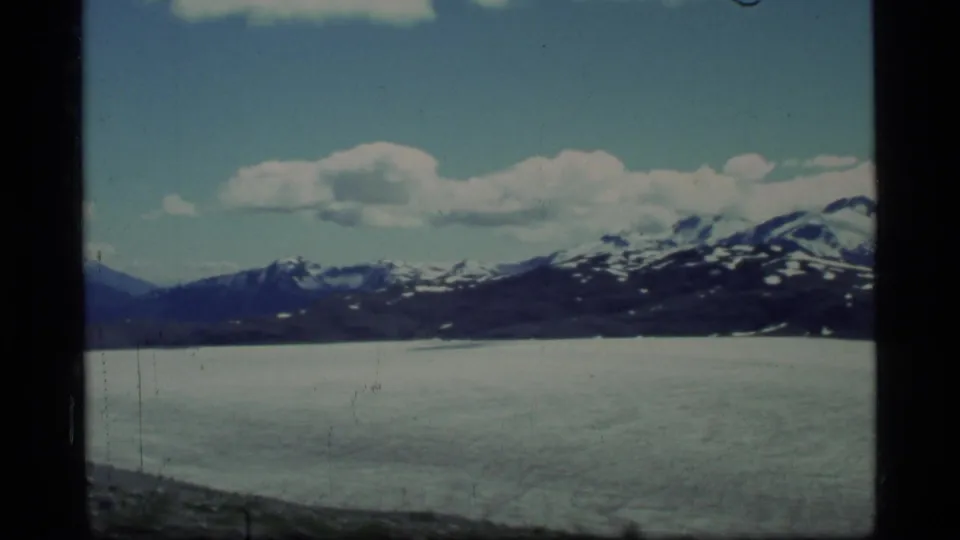 1977 Glacier Bay 8mm frame with a wide icy expanse, distant snow-capped mountain range, and clouds across a blue sky.