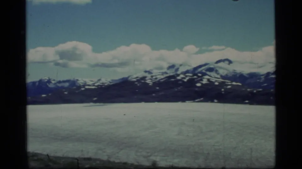 Glacier Bay landscape in 1977 8mm footage, with an icy foreground, distant dark mountains, snow patches, and drifting clouds.