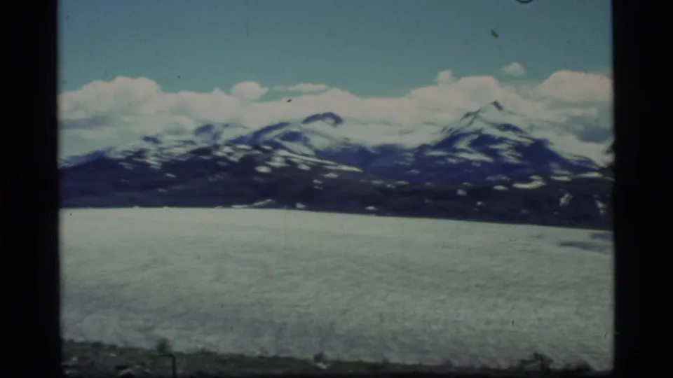 1977 Glacier Bay 8mm frame with a pale glacier foreground, dark mountain ridge, snow patches, and clouds across the horizon.