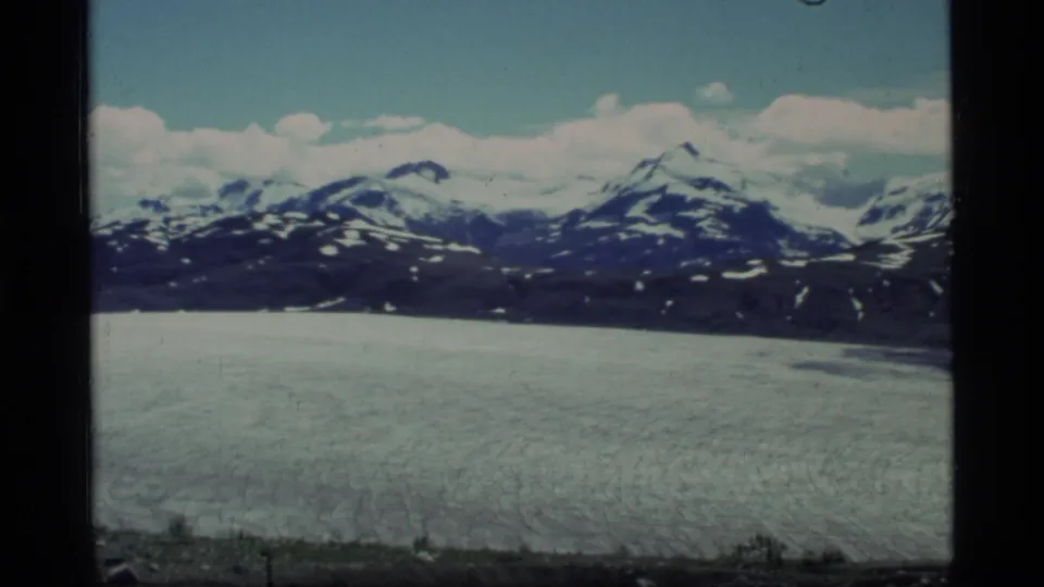 Glacier Bay, Alaska frame from 1977 showing textured glacier ice below a dark ridge and snow-covered mountain peaks.