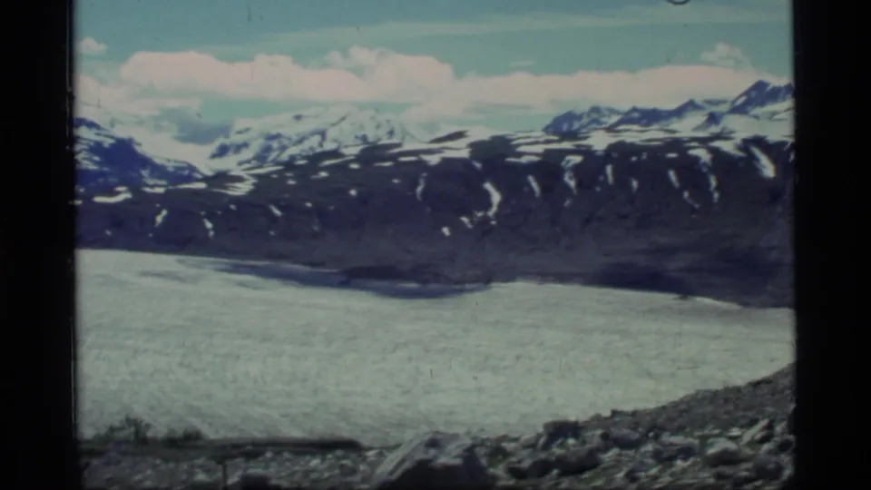 1977 Glacier Bay home-movie frame showing rocky foreground beside glacier ice with snow-marked mountains under a bright sky.