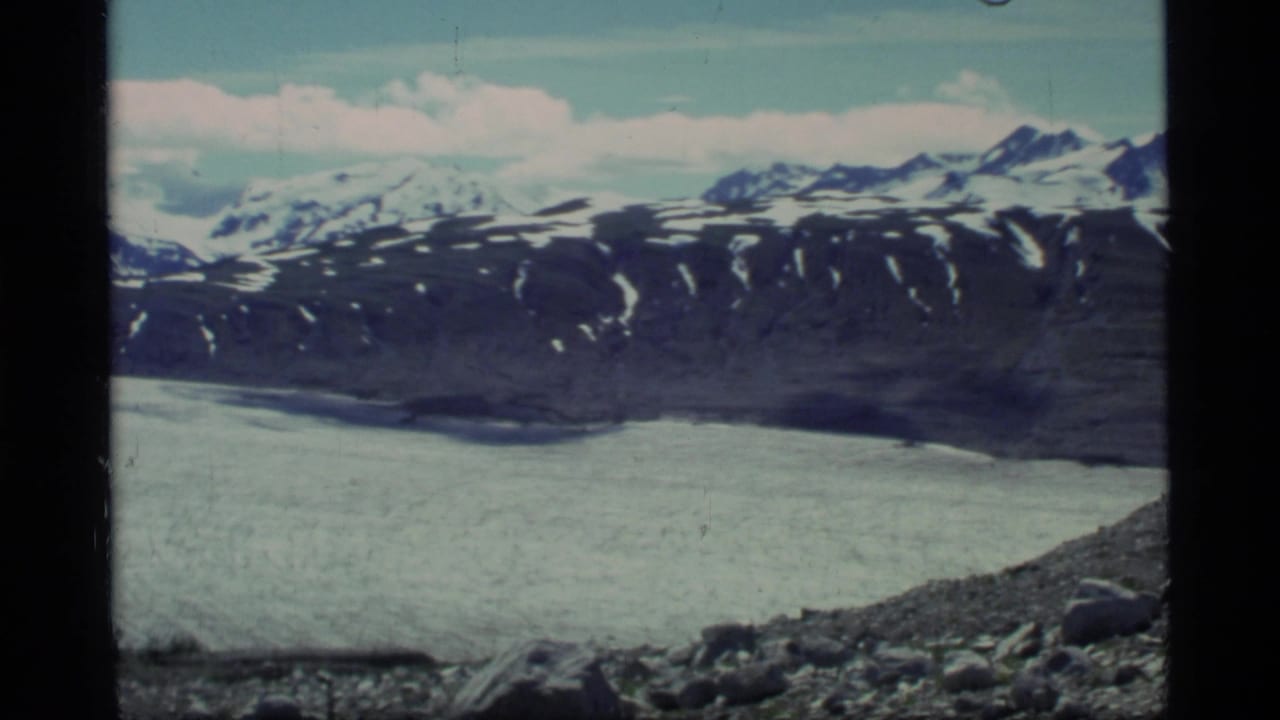 Glacier and snow-capped mountain range seen from a rocky slope.