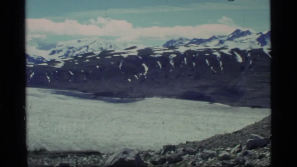 1977 Glacier Bay 8mm frame showing broad glacier ice below dark ridges and snow-capped mountains under pale clouds.