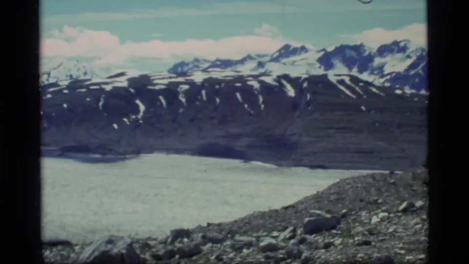 1977 Glacier Bay home-movie frame with glacier ice, rocky foreground, and distant snow-covered peaks in soft 8mm color.