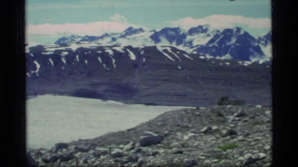 1977 Glacier Bay frame showing a rocky slope above pale glacier ice and a snow-streaked mountain range.
