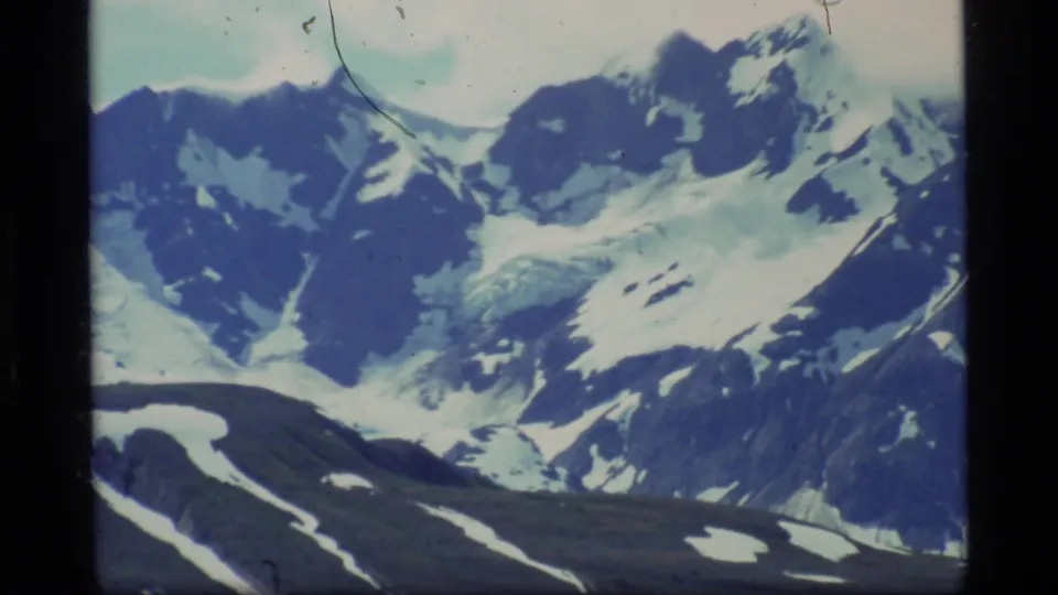1977 Glacier Bay 8mm frame with closer snow-covered mountain peaks, glacier forms, and dark exposed slopes.