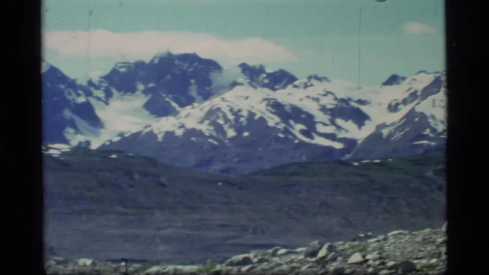 Wide 1977 8mm view of Glacier Bay, Alaska mountains, with snow-covered peaks beyond rocky foreground ground and pale sky.