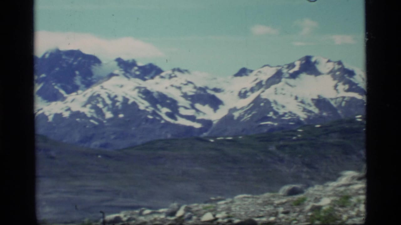 1977 8mm Glacier Bay view of snow-capped mountains with dark ridges and a rocky foreground slope.