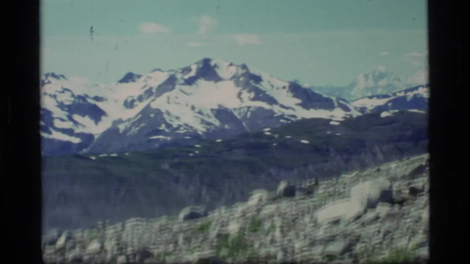 1977 Glacier Bay home-movie frame showing a rocky alpine slope, boulders, and distant snow-covered mountains.
