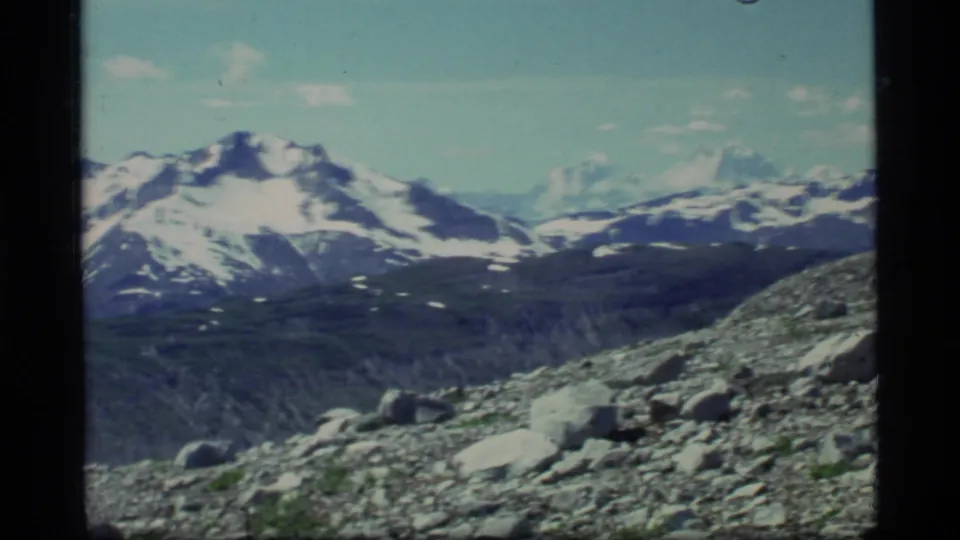 1977 8mm frame from Glacier Bay with a snow-covered peak, scree slope, scattered rocks, and sparse green vegetation.