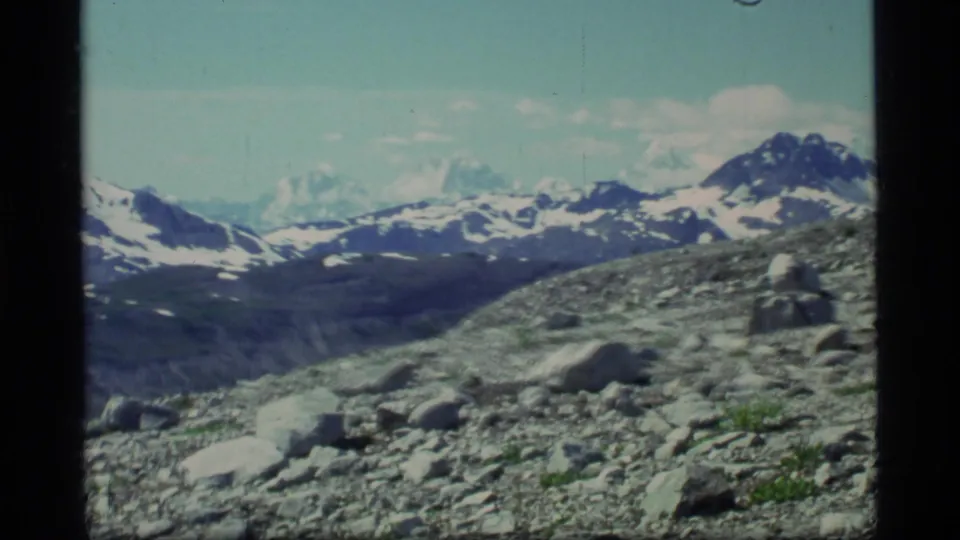 1977 Glacier Bay landscape frame showing a rocky slope in the foreground and snow-streaked mountains under clear sky.