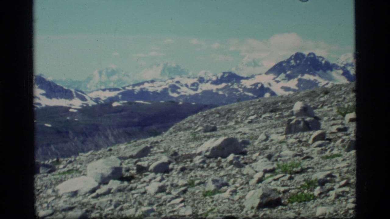 1977 8mm Glacier Bay view across rocky ground toward distant snow-capped mountain peaks and scattered clouds.