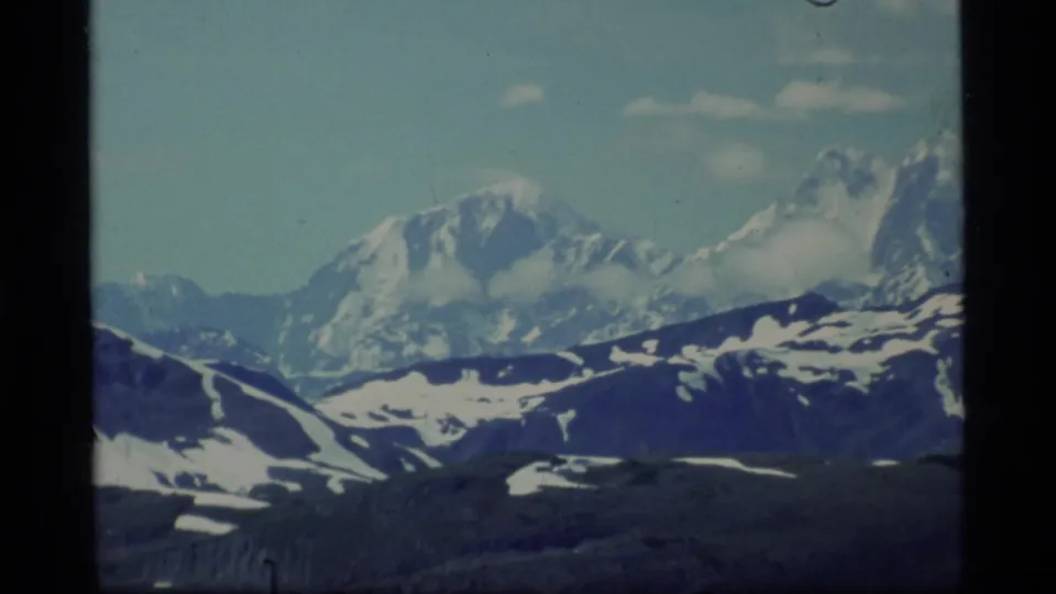 1977 8mm Glacier Bay mountain view with glaciated peaks, snow-streaked ridges, clouds, and visible frame edges.