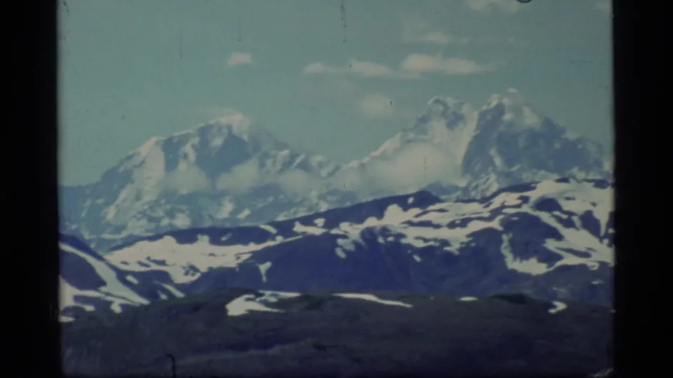 1977 8mm Glacier Bay frame showing high snow-capped peaks, dark foreground ridges, and muted home-movie color.