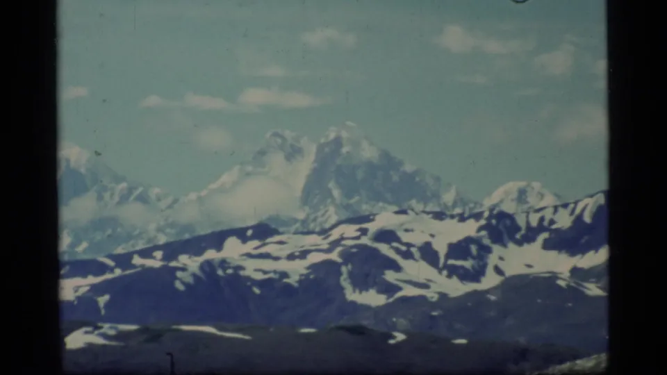 1977 8mm Glacier Bay view of a distant glaciated peak, snow-streaked ridges, pale sky, and dark film edges.