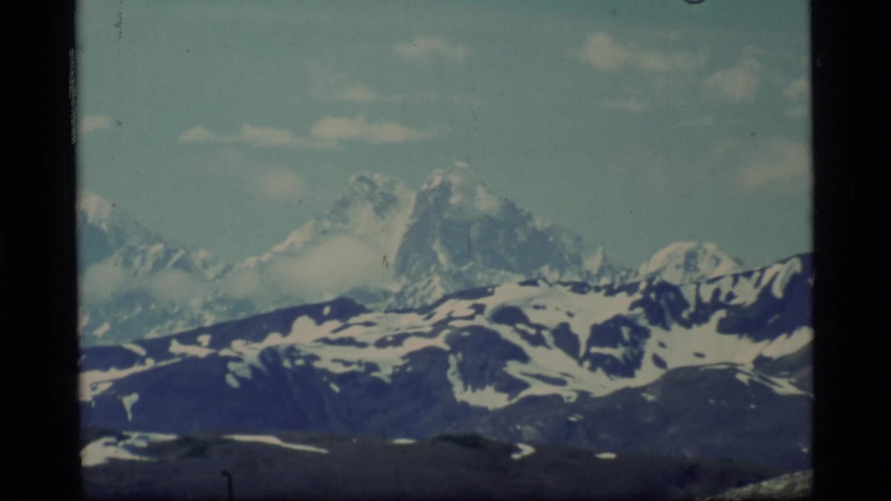1977 Glacier Bay home-movie frame showing distant snow-covered mountains beneath pale blue sky and clouds.