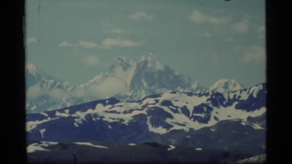 1977 Glacier Bay home-movie frame showing distant snow-covered mountains beneath pale blue sky and clouds.
