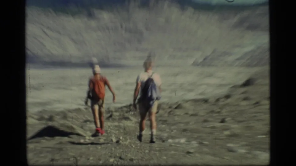 Two hikers in shorts and backpacks cross rocky ground in Glacier Bay, Alaska, with glacial ice and mountains ahead.