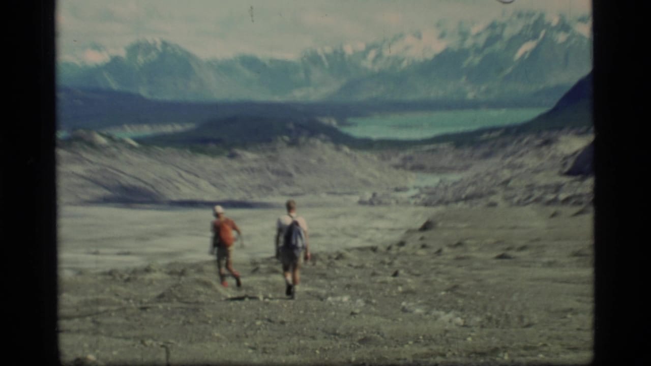 Two hikers walk across rocky glacial terrain with mountains and a lake beyond.