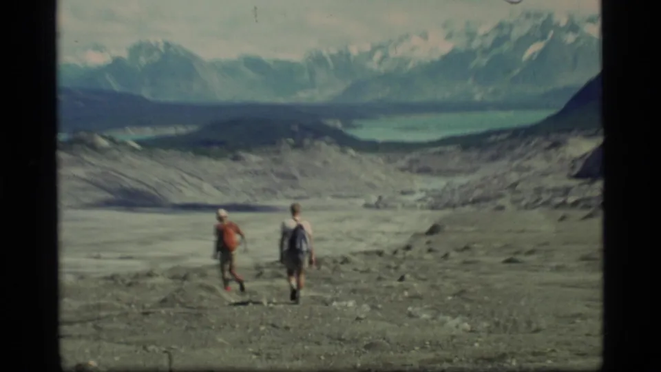 Two hikers in Glacier Bay, Alaska, walk over a barren rocky glacial landscape with a pale lake and snow-capped mountains in the distance.