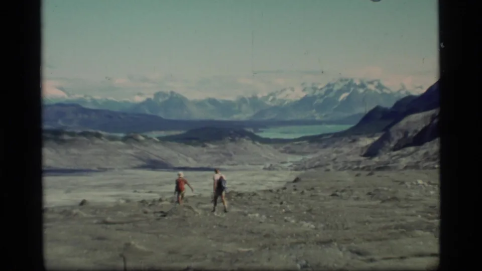 In the 1977 Glacier Bay footage, two hikers continue across open glacial terrain below distant snow-capped mountains and a turquoise lake.