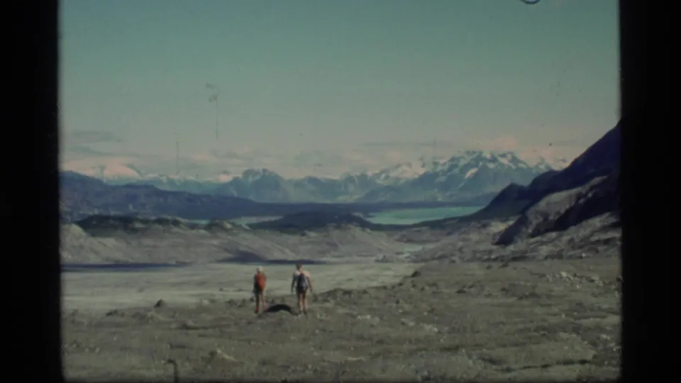 Two distant hikers appear small against the rocky Glacier Bay landscape, with a pale lake, dark slopes and snow-capped mountains beyond.