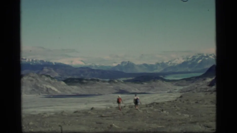 Two hikers, seen at a distance in Glacier Bay, cross rocky glacial ground beneath a broad sky with a lake and snow-capped mountains behind them.