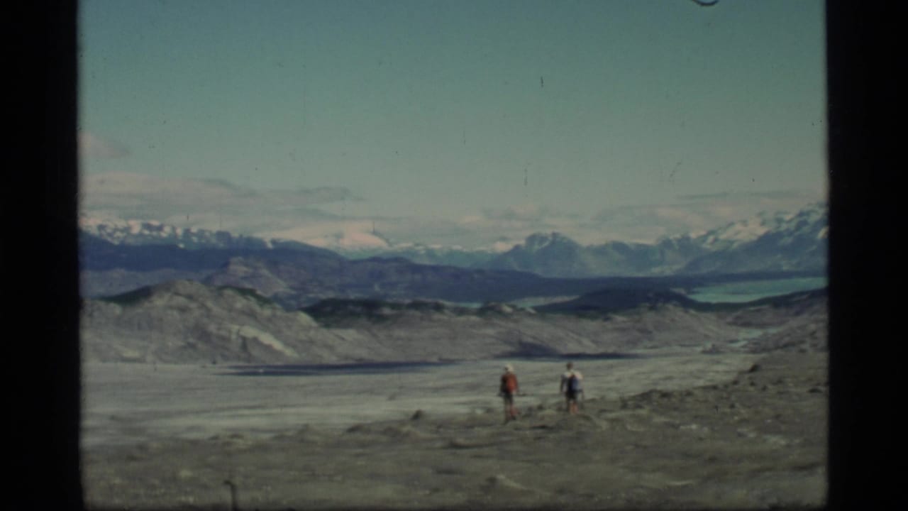 Two backpacked hikers cross a rocky glacial landscape with mountains in the distance.