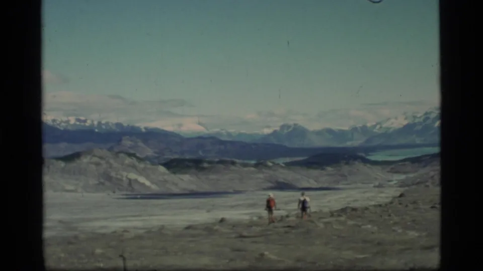 Two hikers with backpacks traverse open glacial terrain at Glacier Bay, Alaska, in 1977 home-movie footage.