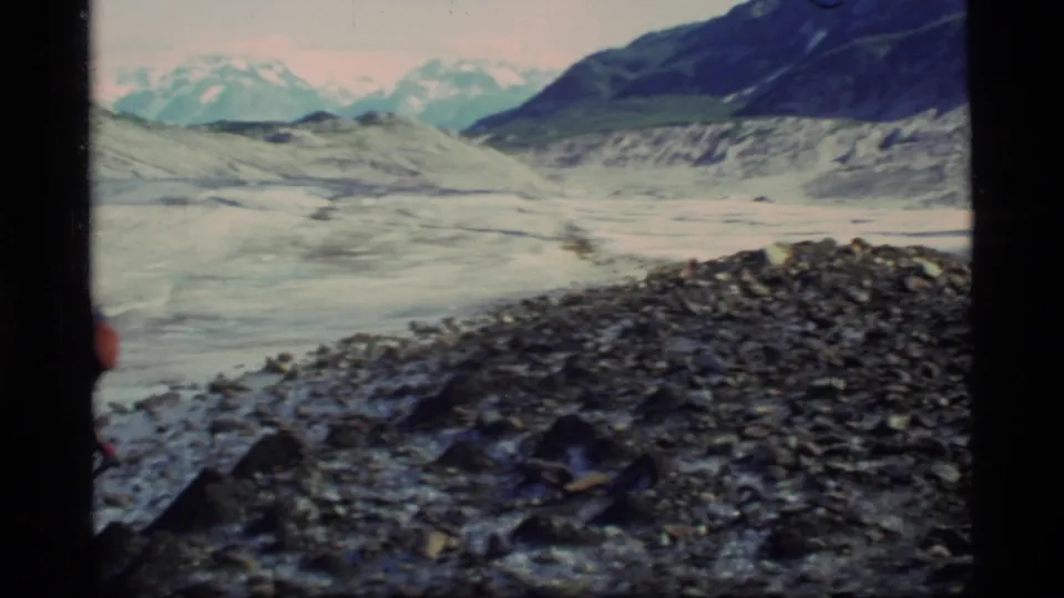 A Glacier Bay rocky moraine and pale glacial ice fill the frame, with mountain slopes beyond.