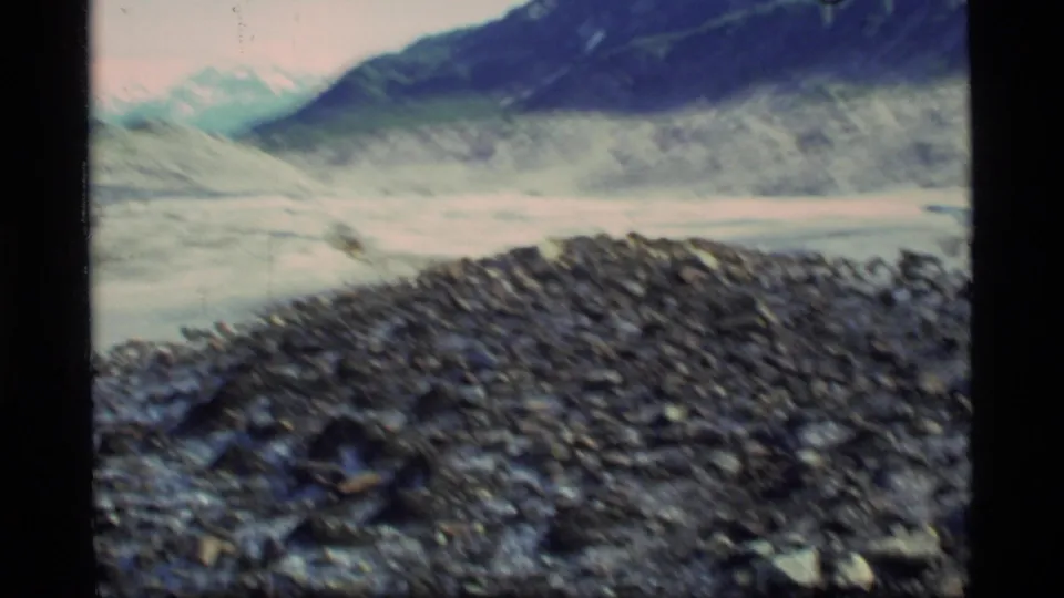 Handheld 8mm footage looks across a blurred rocky foreground toward glacier ice and dark mountain slopes.