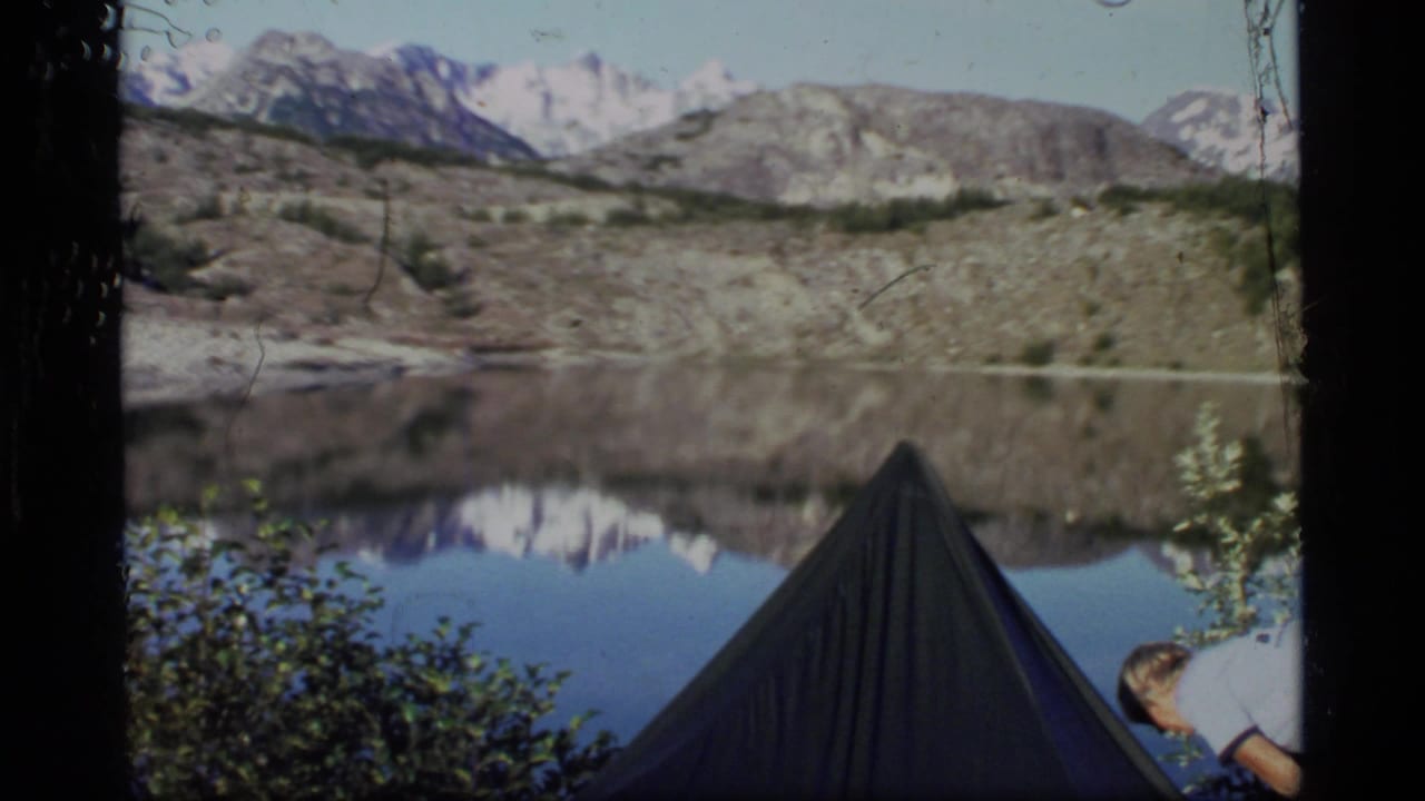 1977 Glacier Bay home-movie frame showing a tent by still water, distant snowy mountains, and a partial camper near the foreground.