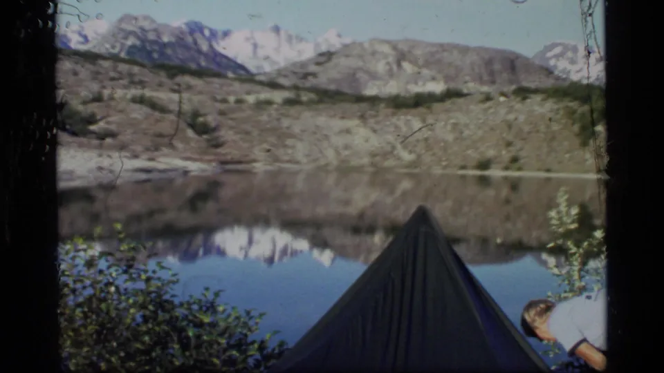 1977 Glacier Bay home-movie frame showing a tent by still water, distant snowy mountains, and a partial camper near the foreground.