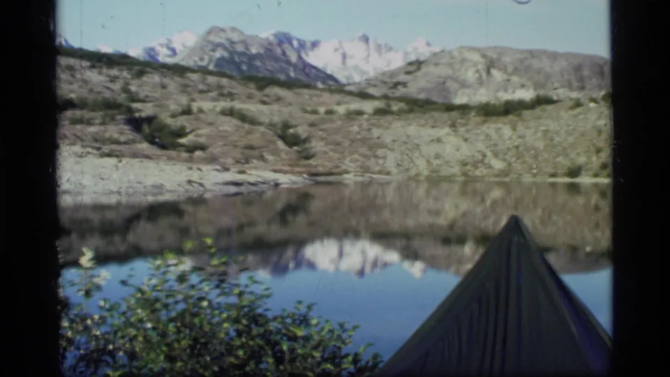 1977 Glacier Bay frame with a tent peak in the foreground, shrubs, still lake water, and snow-capped mountains reflected beyond.