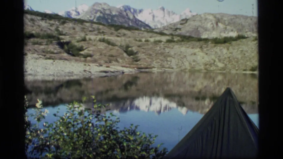 1977 Glacier Bay home-movie view across a calm lake, with shrubs, a tent edge, rocky slopes, and reflected snowy mountains.