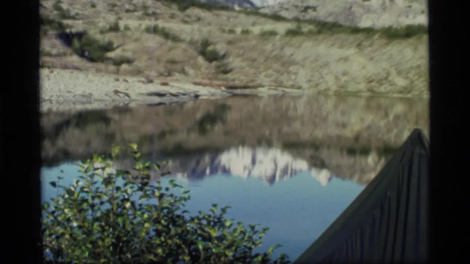 1977 Glacier Bay frame showing the tent edge at lower right, shrubs, still water, and mountain reflections under a pale sky.