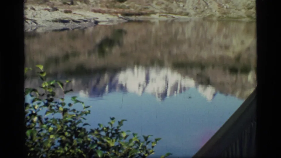 Glacier Bay lake view on 8mm film, with snowy mountain reflections, leafy foreground growth, and a railing at the right edge.