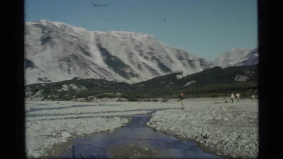1977 Glacier Bay frame showing visitors on a broad rocky riverbed beneath a snow-streaked mountain range and clear sky.