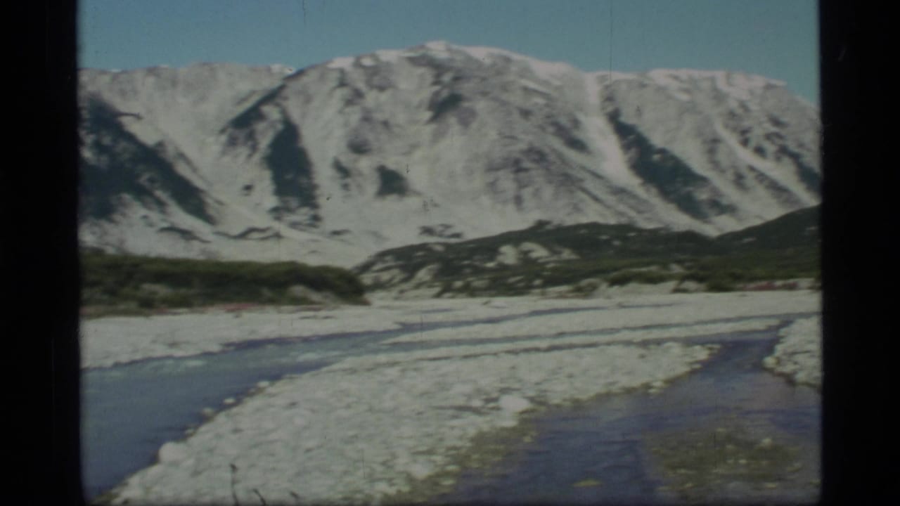 1977 Glacier Bay, Alaska 8mm frame showing a shallow rocky river below snow-capped mountains and low dark vegetation.