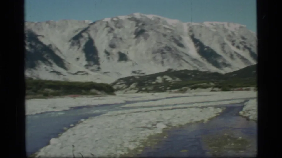 1977 Glacier Bay, Alaska 8mm frame showing a shallow rocky river below snow-capped mountains and low dark vegetation.