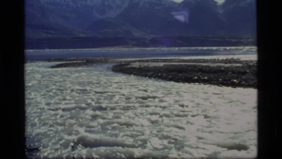 1977 Glacier Bay, Alaska frame showing bright rippled water, a dark gravel bar, and mountains across the valley.